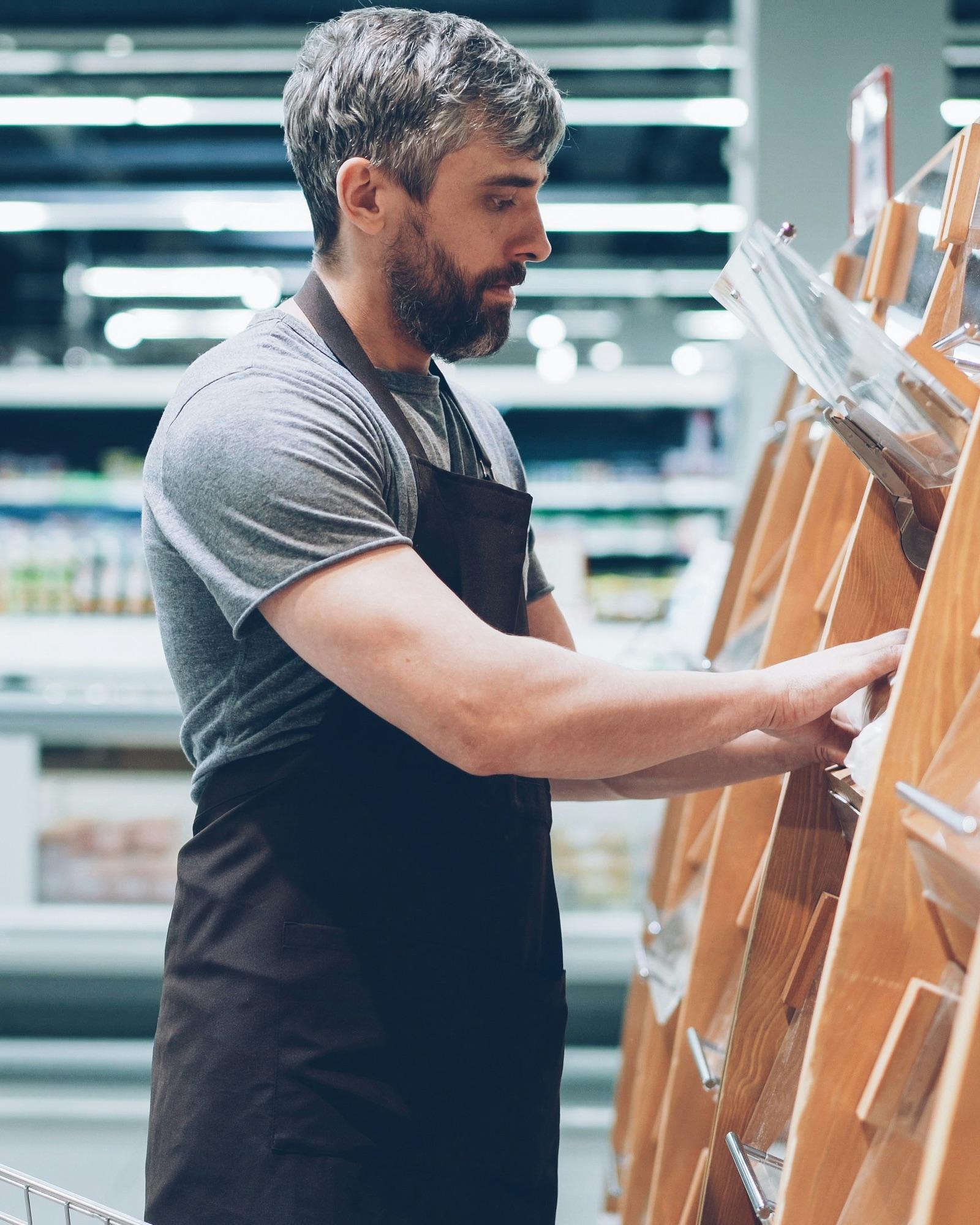 Mitarbeiter im Supermarkt mit dunkler Schürze räumt ein Brotregal ein