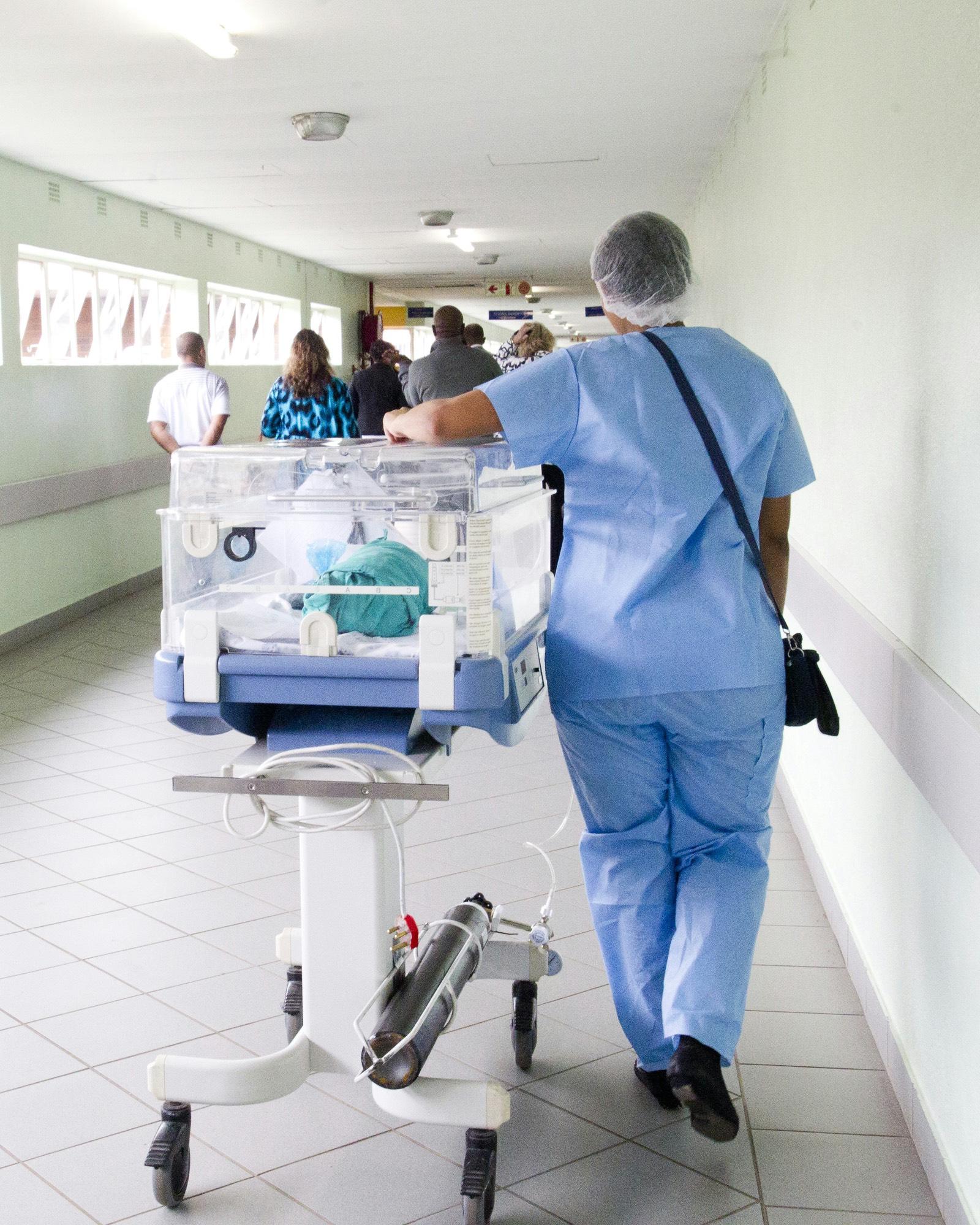 Pflegekraft in blauer Dienstkleidung schiebt einen Inkubator durch einen hellen Klinikflur